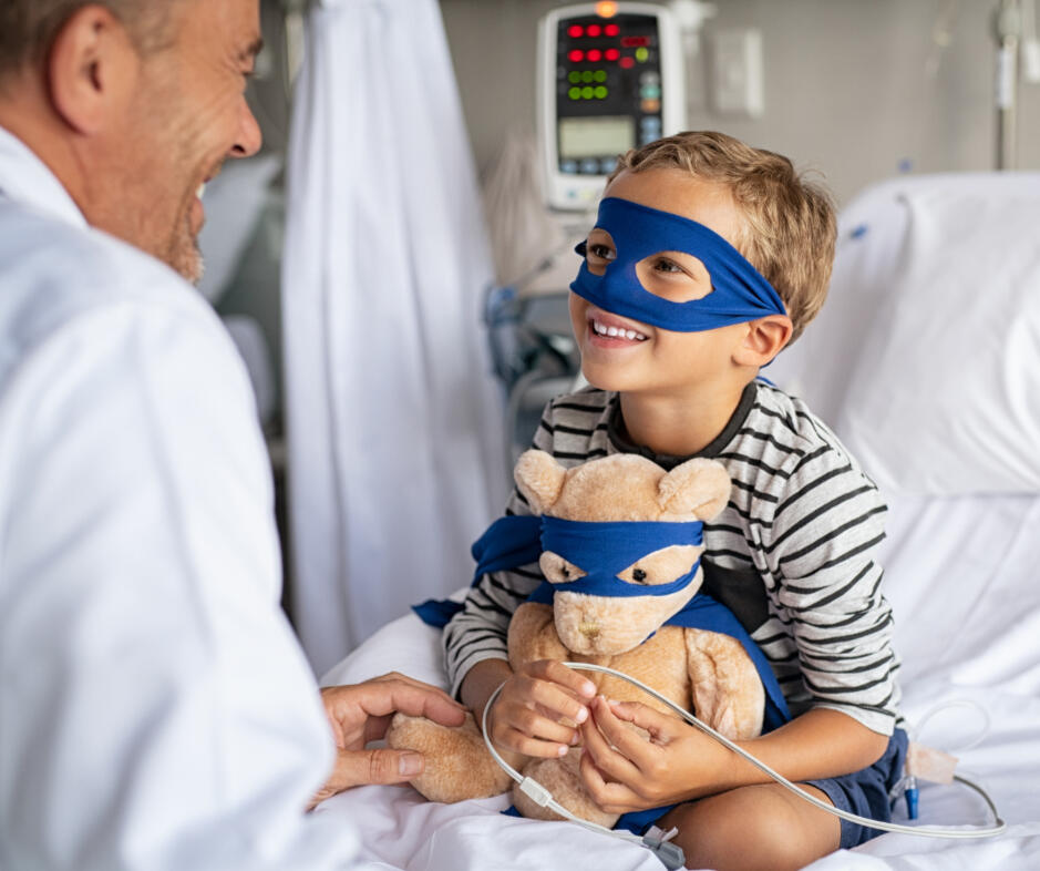 A little boy, wearing a mask, hugs his stuffed bear in a hospital room, symbolizing the emotional support needed during a child' alt="Child patient in hospital bed, masked, embracing a teddy bear also wearing a mask. This image represents the resilience of children and the crucial need for support during extended hospital stays."